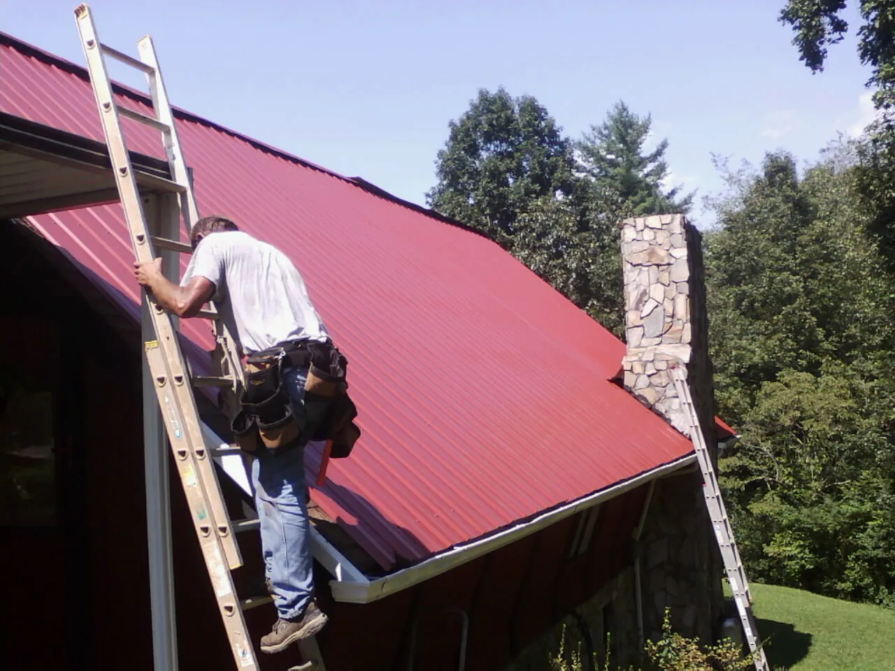 Roofer on a ladder beside a red metal roof and stone chimney — Soffit & Fascia Repair by Roofing Brothers in Fort Lee
